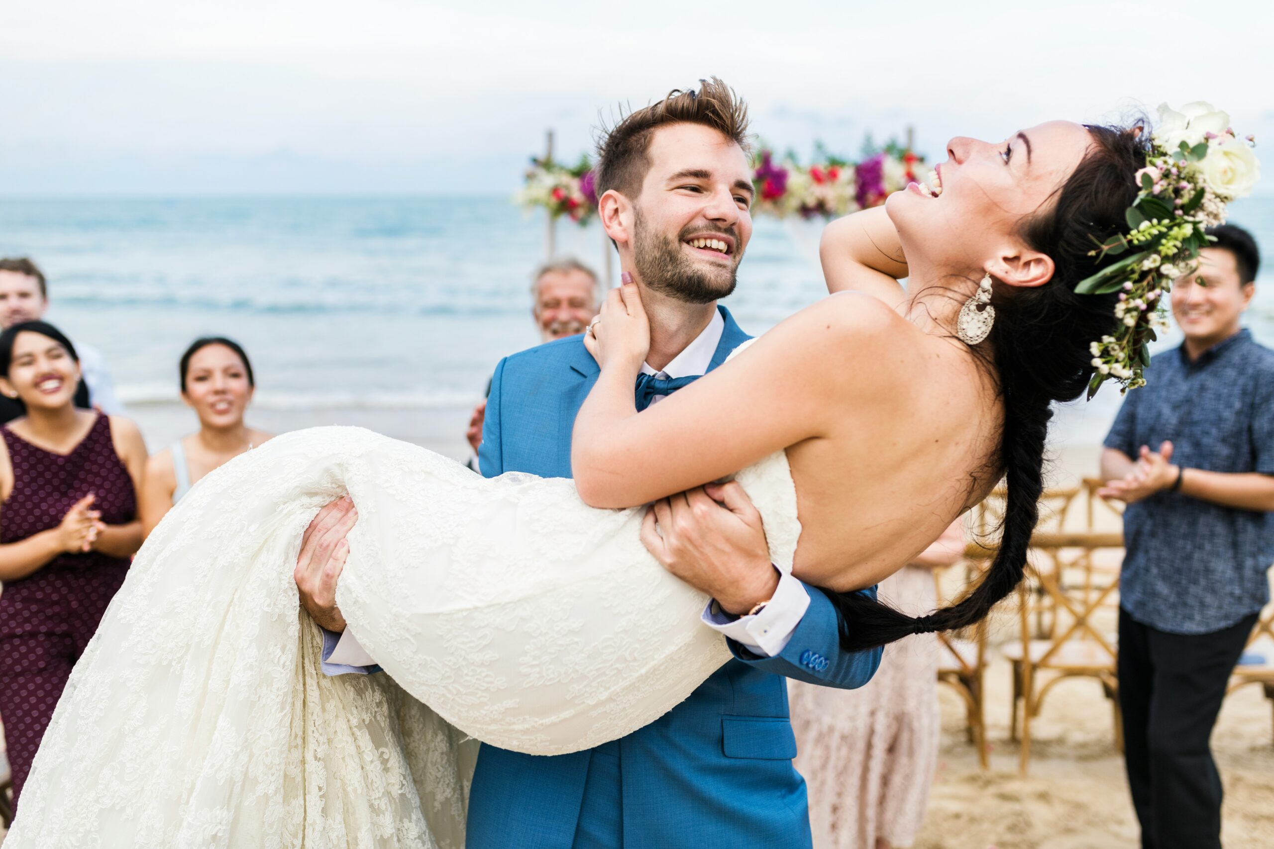 A groom lifts his bride at an oceanfront wedding ceremony as guests applaud