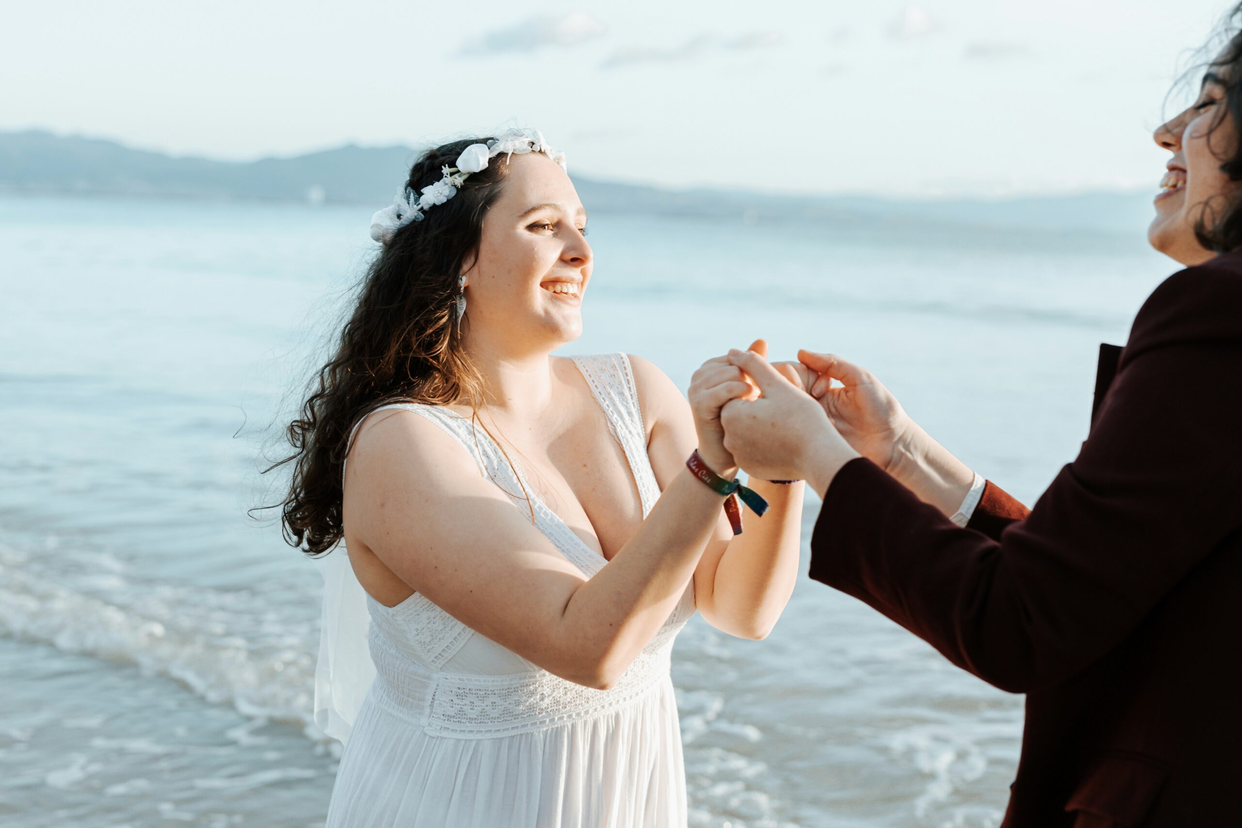 A bride in a lace gown holds hands with a guest at the water's edge