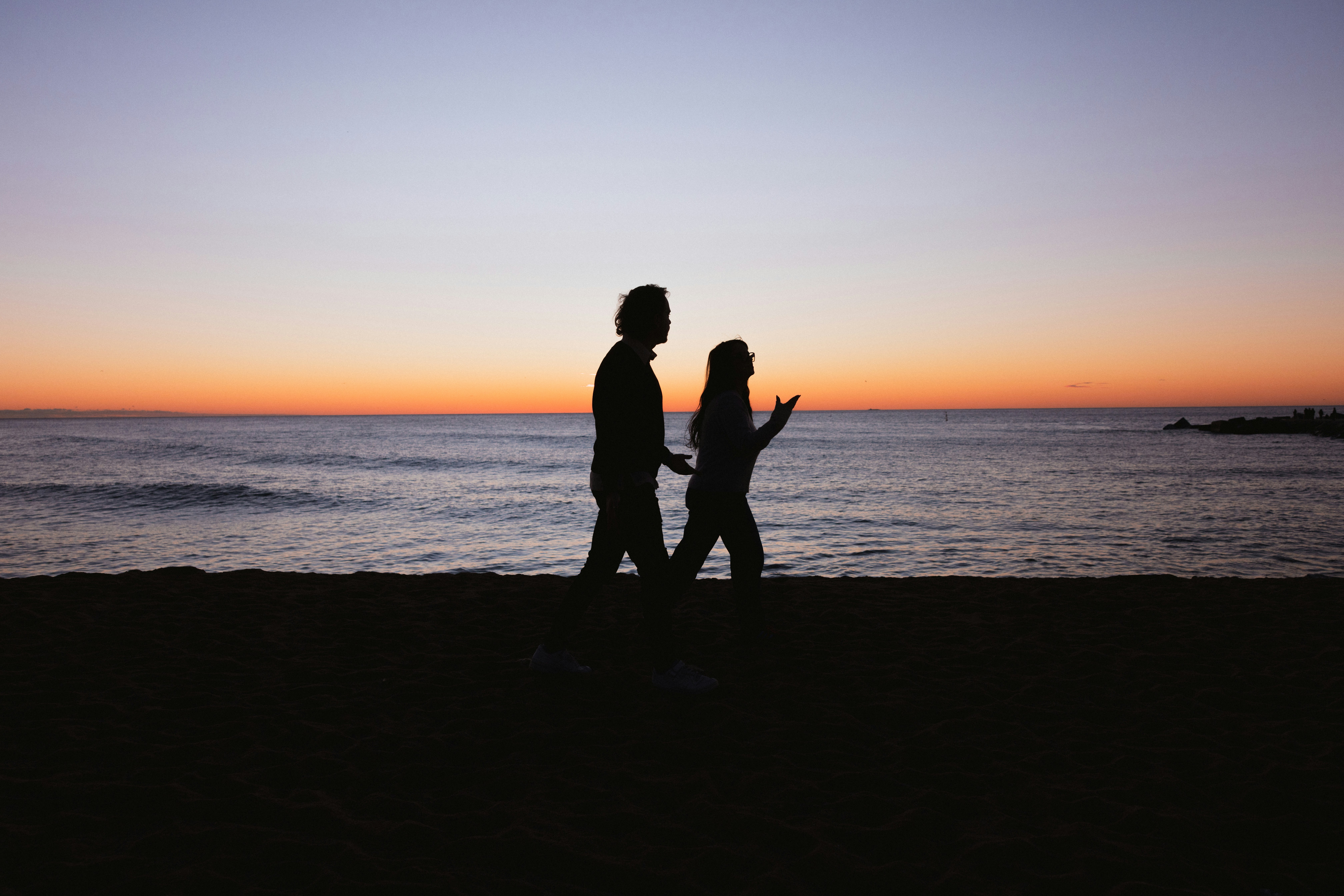 A couple walk hand in hand along the shoreline silhouetted against a sunset