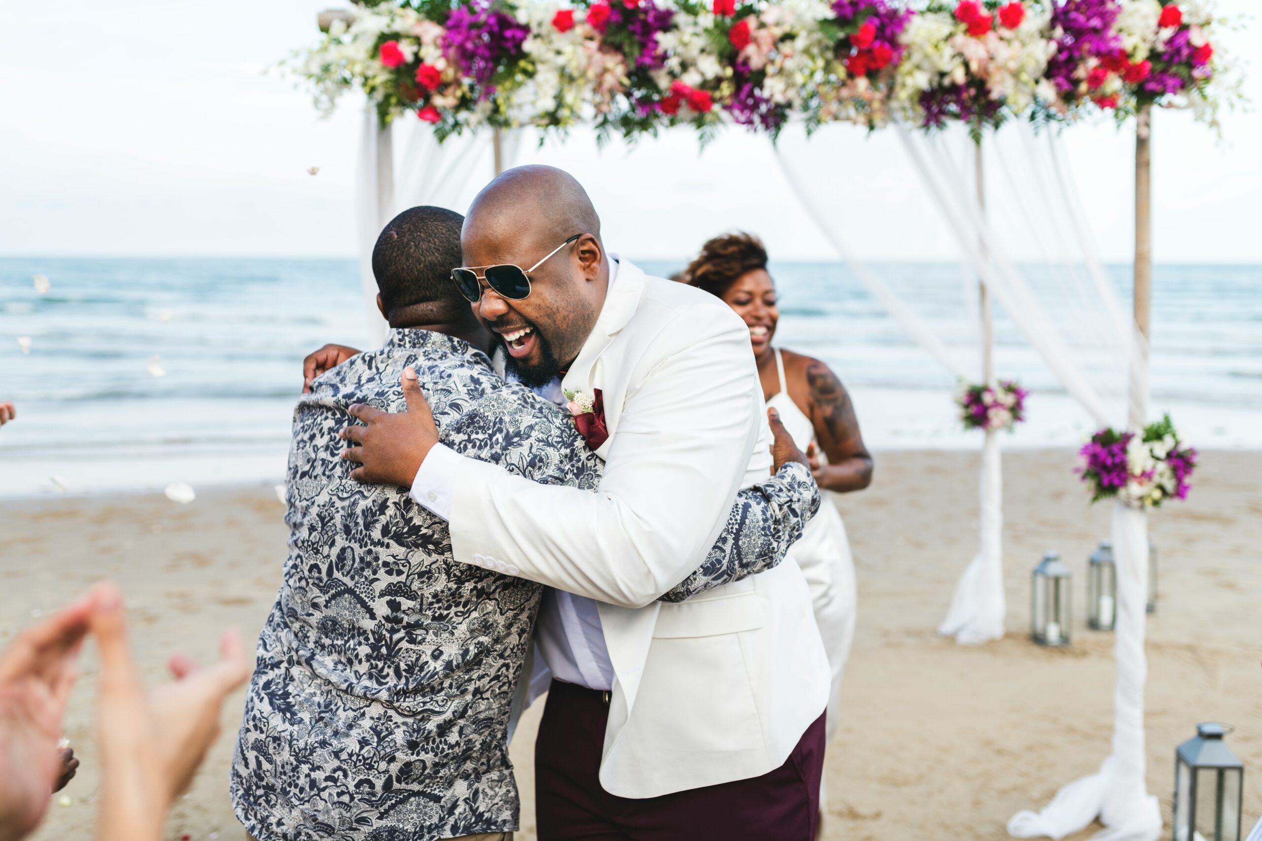 A groom embraces a guest under a floral arch at an oceanfront wedding ceremony