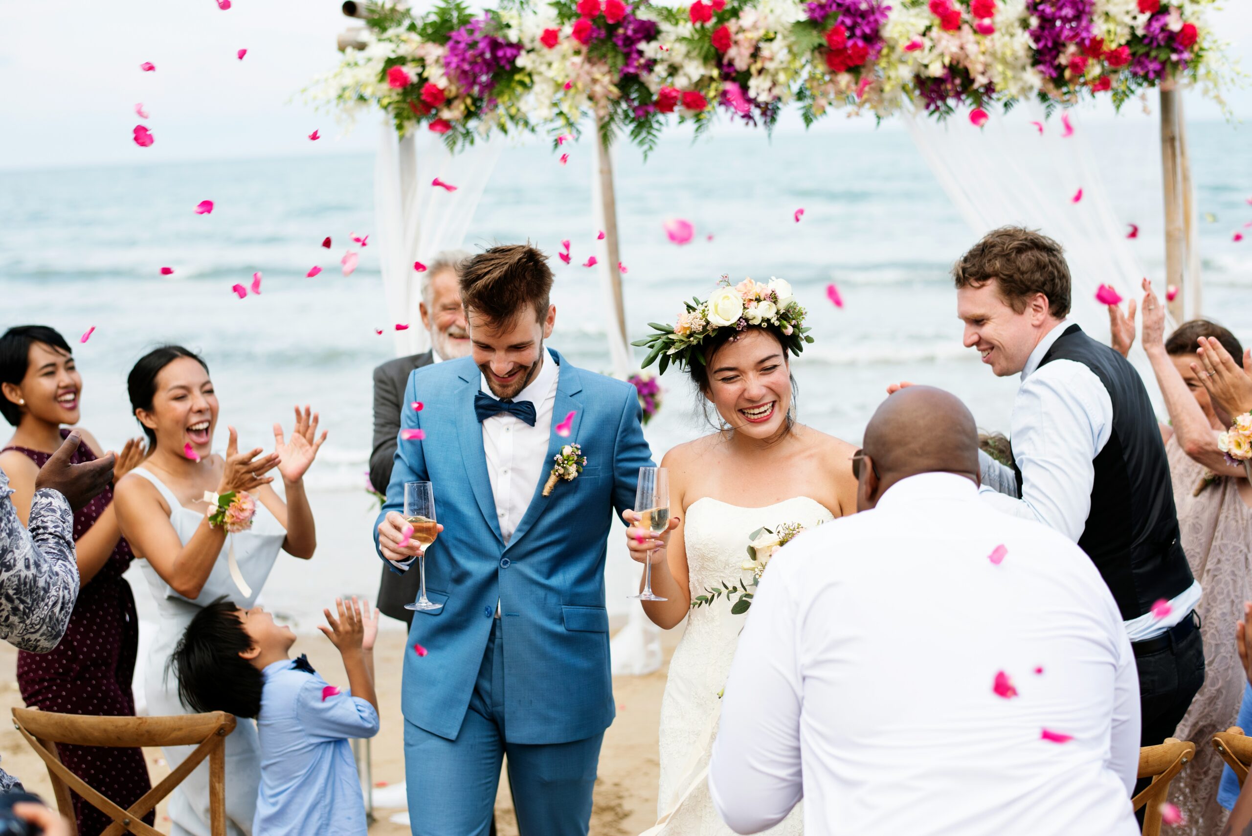 Guests throw flower petals over a couple standing beneath a floral arch by the ocean